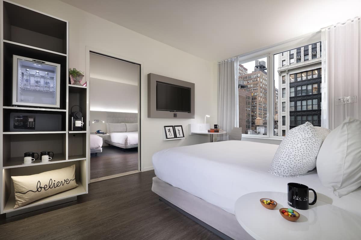A modern hotel room with a large bed, a TV mounted on the wall, a built-in shelf with a refrigerator, and a window offering a city view.