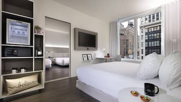 A modern hotel room with a large bed, a TV mounted on the wall, a built-in shelf with a refrigerator, and a window offering a city view.