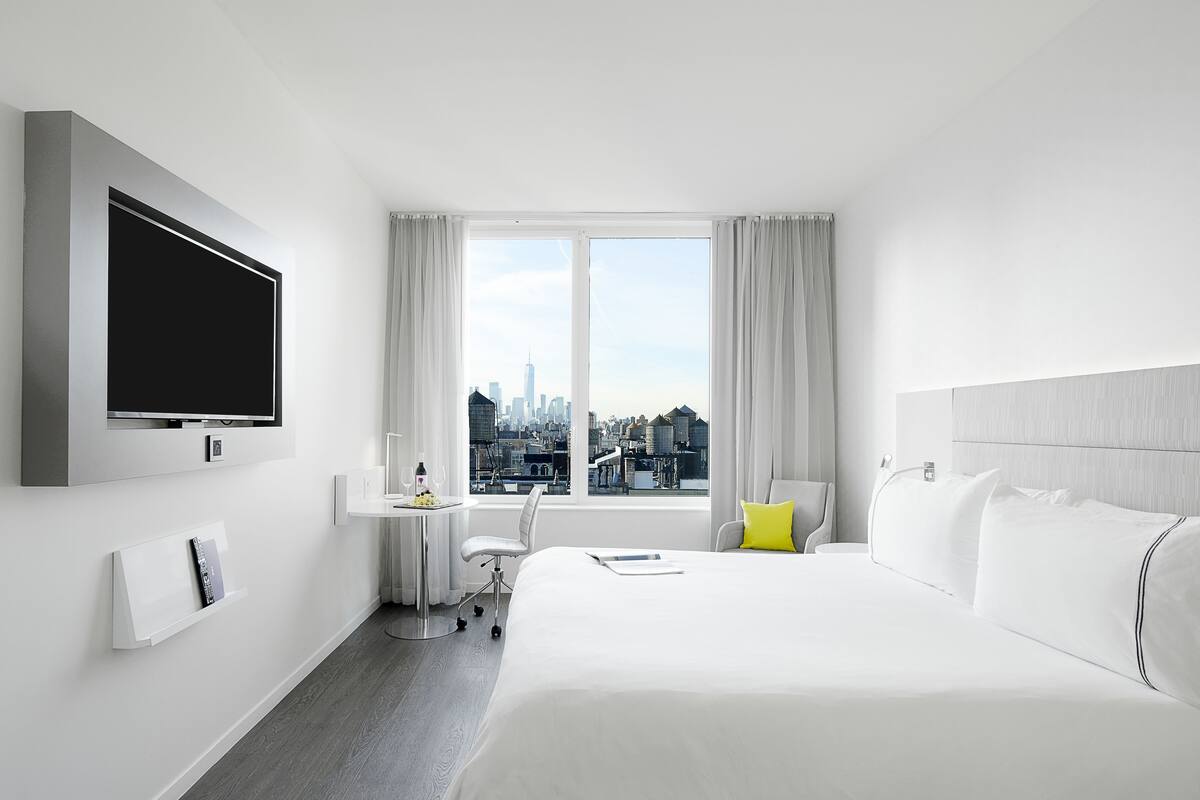 A hotel room with a large bed, a desk with a chair, a flat-screen TV mounted on the wall, and a window with a city view.