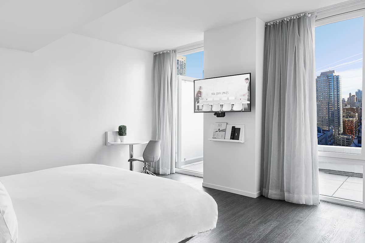 A modern hotel room with a large bed, a TV mounted on the wall, a small table with a plant, and a city view through the window.