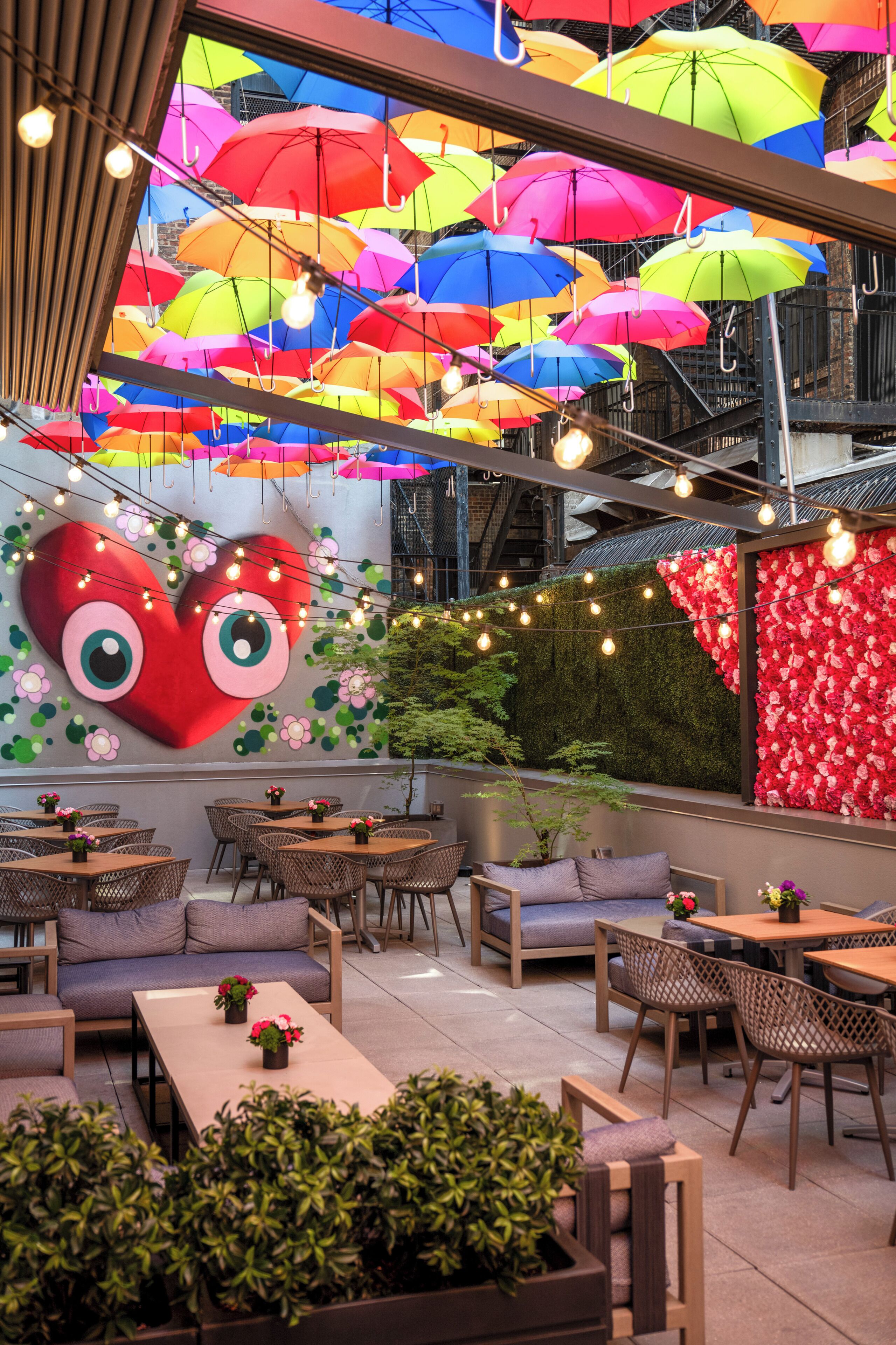 Uma área de refeições no terraço com várias mesas e cadeiras, um grande mural em formato de coração e guarda-sóis coloridos por cima.