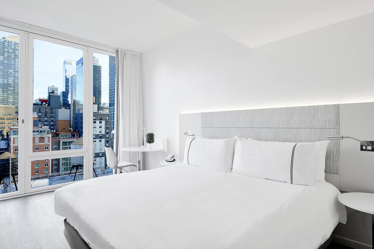 A modern hotel room with a large bed, a bedside table, a small desk, and a city view through floor-to-ceiling windows.