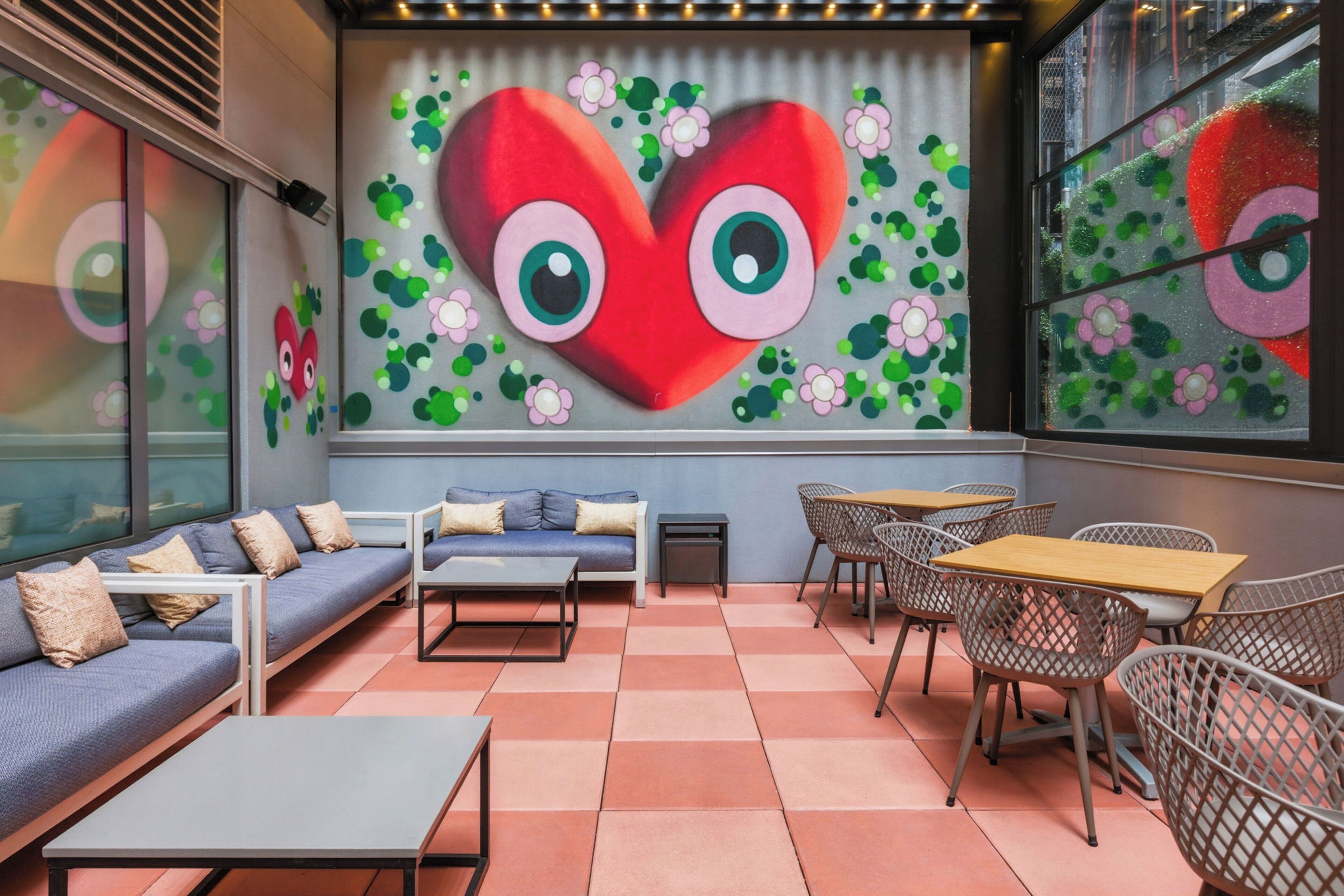 Un salón moderno con sofá, mesas y sillas, un mural grande con forma de corazón y una ventana con vistas.