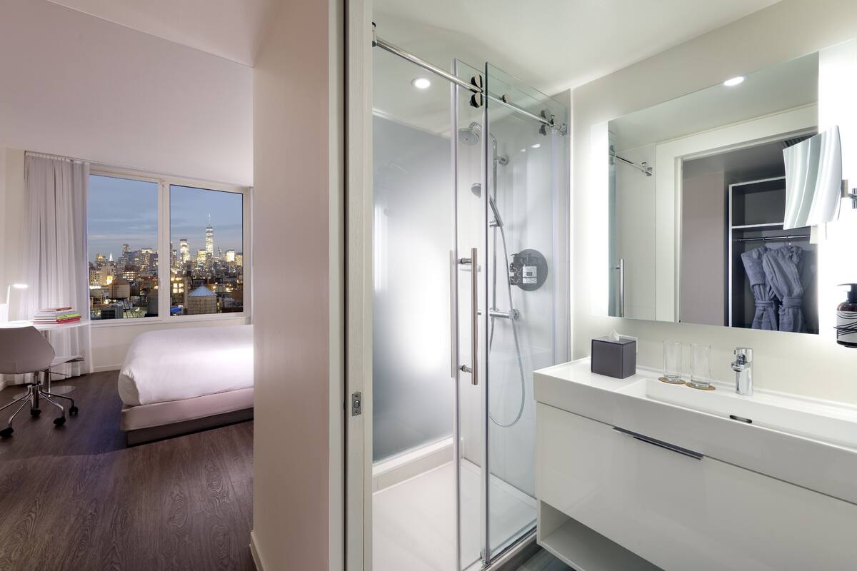 A modern hotel room with a glass-enclosed shower, a large bed, a white vanity with a mirror, and a view of the city skyline.
