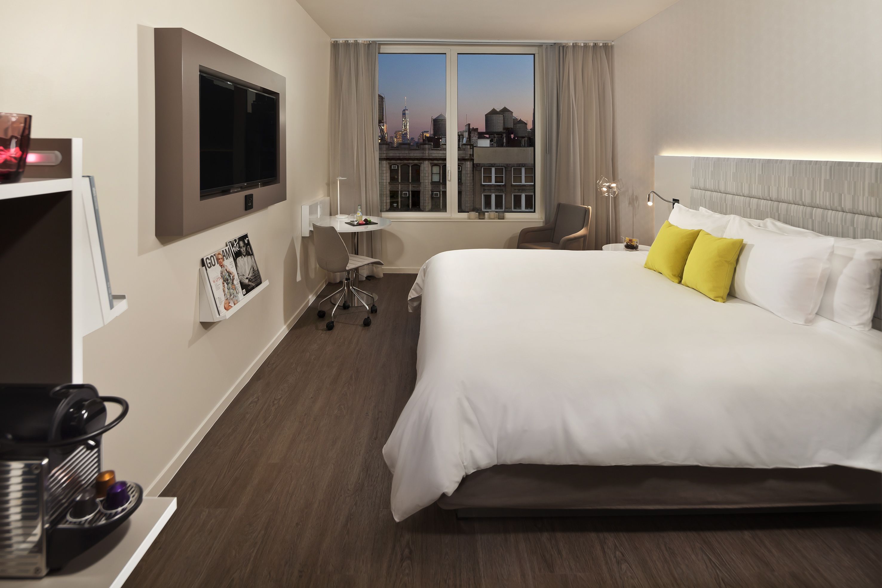 Ein Hotelzimmer mit einem großen Bett, einem Schreibtisch, einem Stuhl, einem an der Wand befestigten Fernseher und Blick auf die Stadtlandschaft.