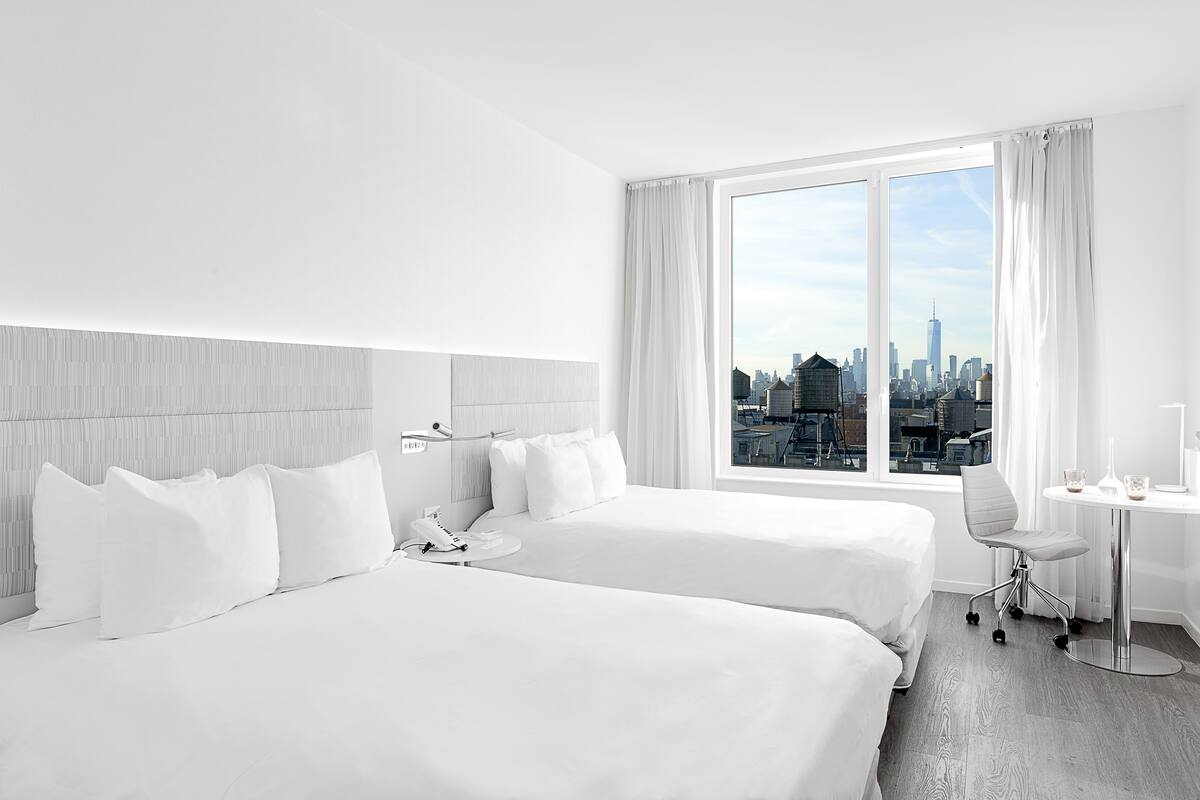 A hotel room with two beds, a desk, a chair, a phone, and a window with a city view.