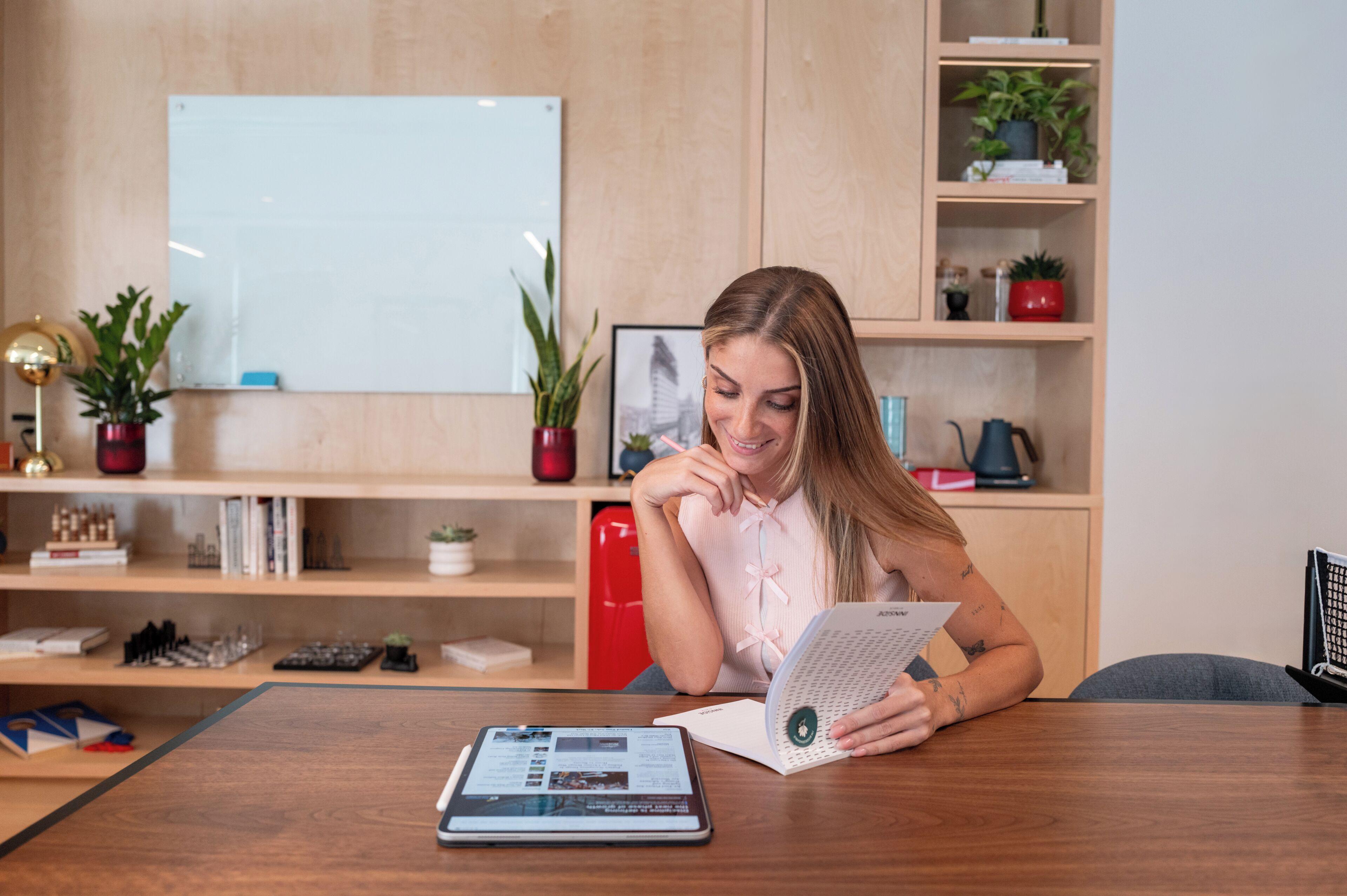 A person sitting at a wooden table with a laptop and a magazine, in a room with shelves and a whiteboard.