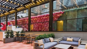 Un café moderno con suelo a cuadros, zona de estar con sofás y mesas, y amplias ventanas con arreglos florales decorativos.