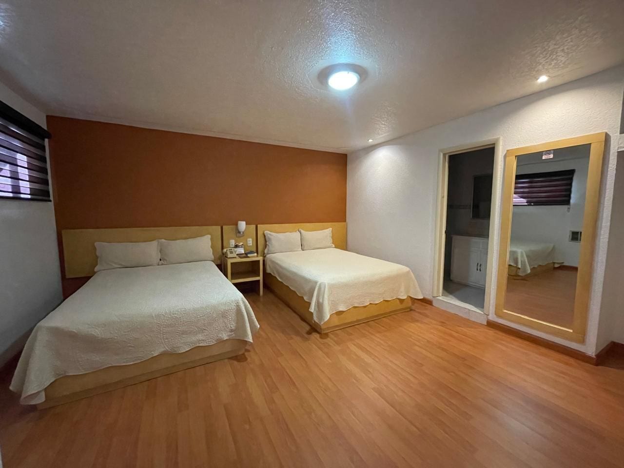 Un dormitorio con dos camas, piso de madera, una mesita de noche, un espejo y una puerta que da a otra habitación.