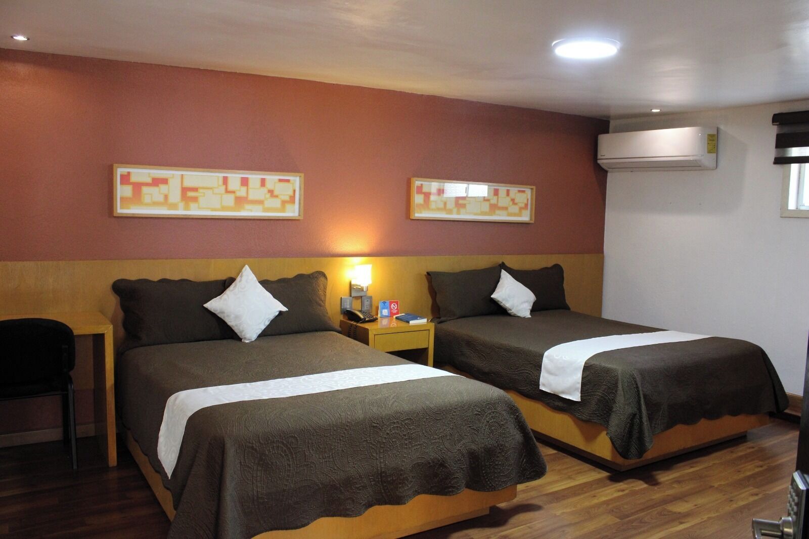 A hotel room with two beds, a nightstand with a lamp, a chair, and an air conditioning unit.