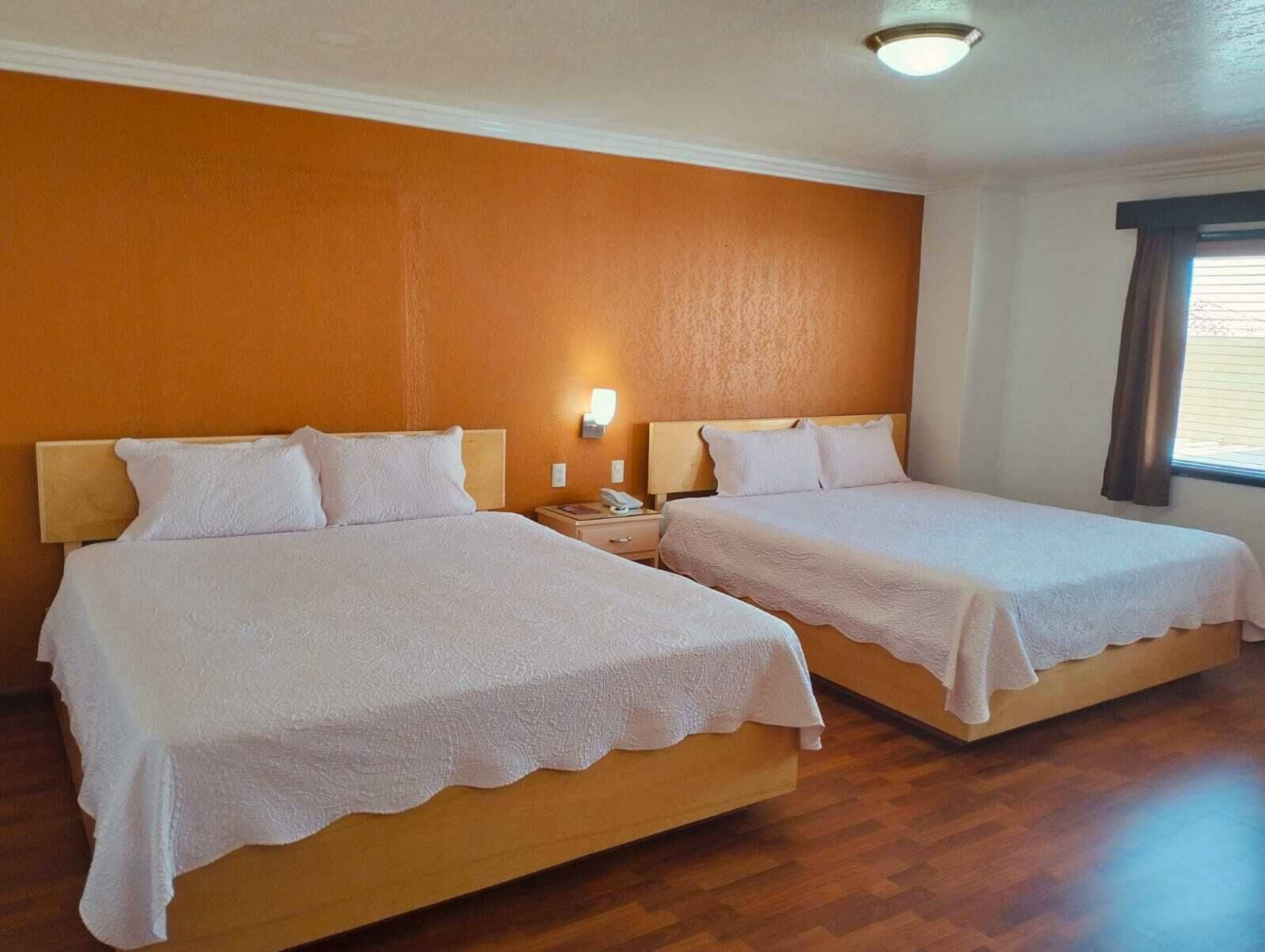 A hotel room with two beds, wooden headboards, a nightstand with a lamp, and a window with curtains.