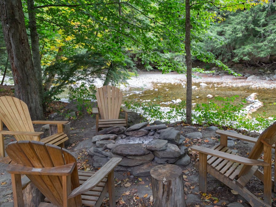 A rustic outdoor seating area with Adirondack chairs and a stone fire pit surrounded by trees and a small stream.