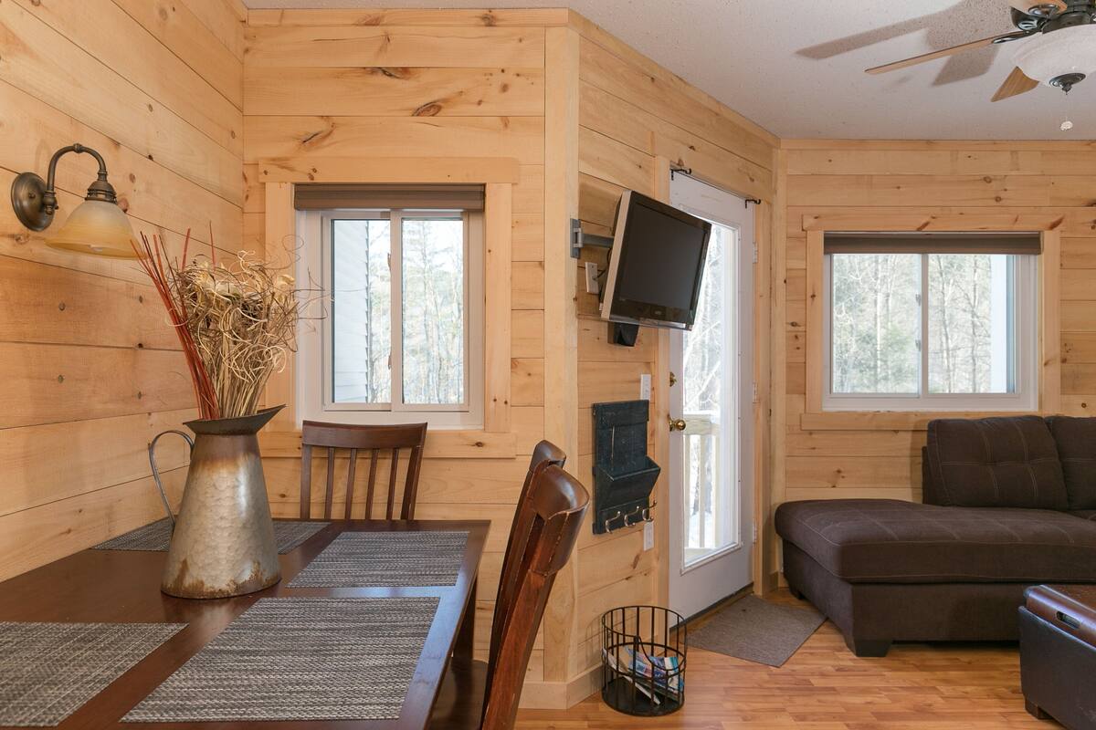 Interior de uma cabana de madeira com uma mesa de jantar, cadeiras, um sofá, uma televisão e uma lareira.