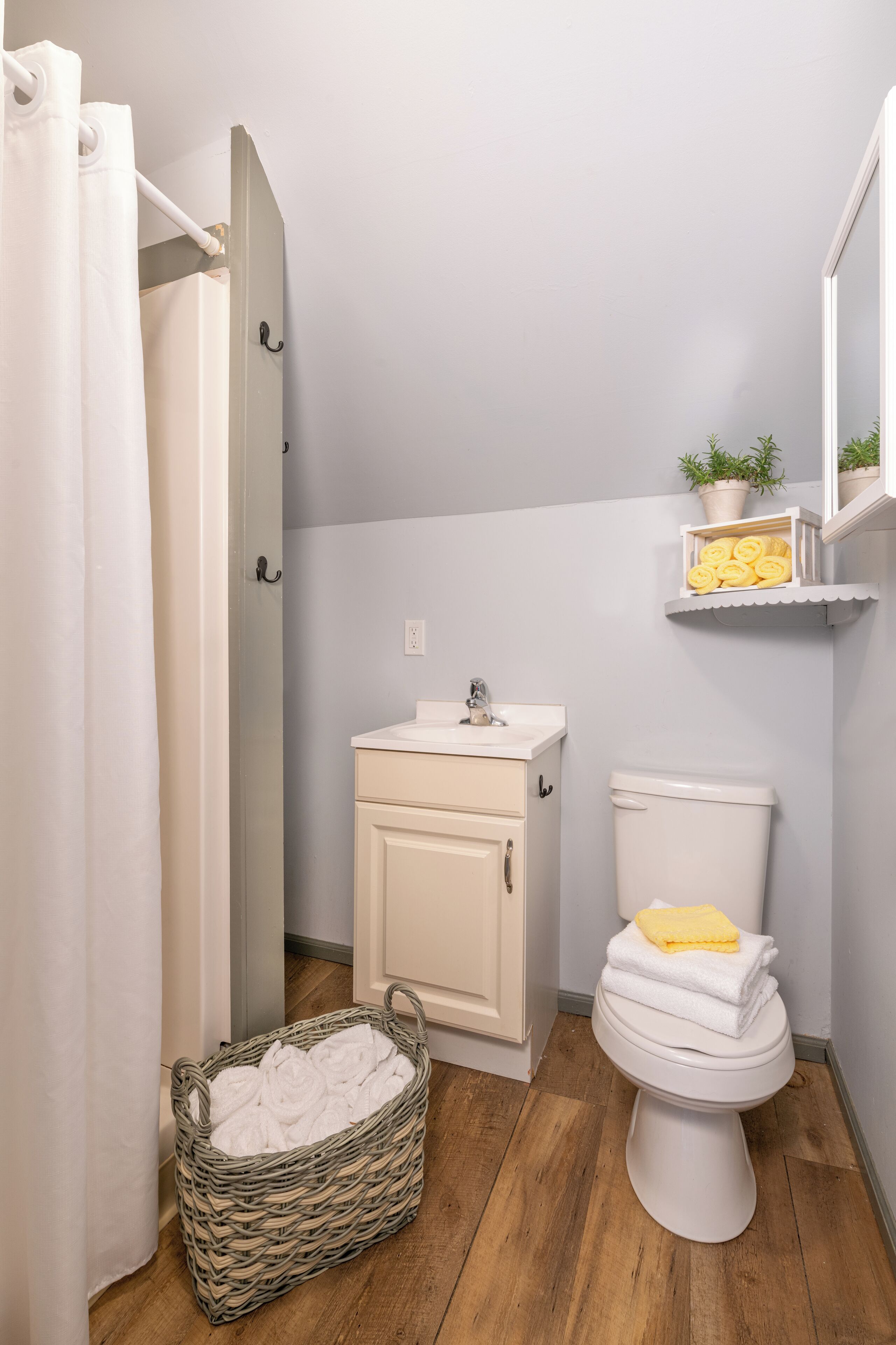 Un baño con un lavabo blanco, un inodoro, una cesta de mimbre con toallas y un estante con plantas en macetas.