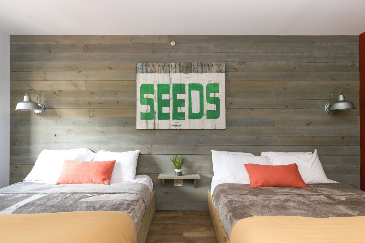 Quarto com duas camas, cada uma com almofadas brancas e laranja, uma mesinha de cabeceira com uma planta, e um letreiro fixado na parede com a palavra "SEEDS".