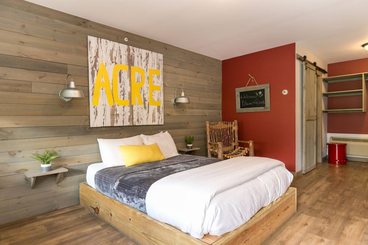 Quarto com cama de madeira, um grande letreiro 'Acre', uma parede vermelha e uma pequena planta num criado-mudo.