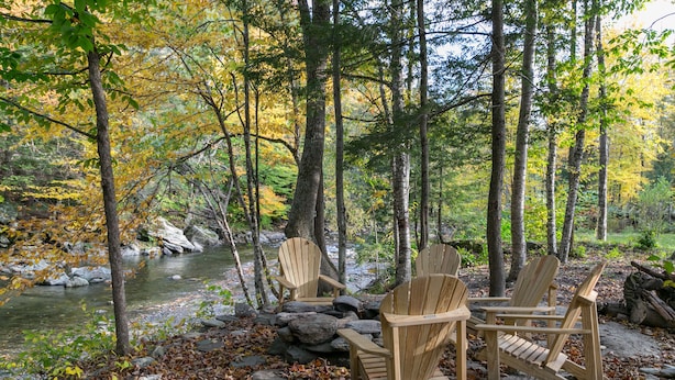 Un área boscosa con un río, rocas y tres sillas de madera tipo Adirondack.