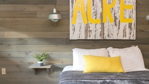 Un dormitorio con pared de madera, una cama con almohadas blancas y amarillas, una mesita de noche pequeña con una planta y una lámpara de pared.