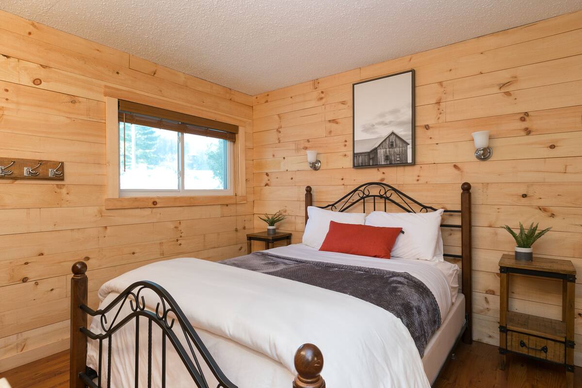 Quarto com painéis de madeira, uma cama, uma janela, um quadro na parede e uma mesa de cabeceira com uma planta.