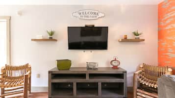 Un salon avec un téléviseur fixé au mur, des chaises en bois, une console ornée d’objets décoratifs et une pancarte indiquant « Bienvenue à la ferme ».