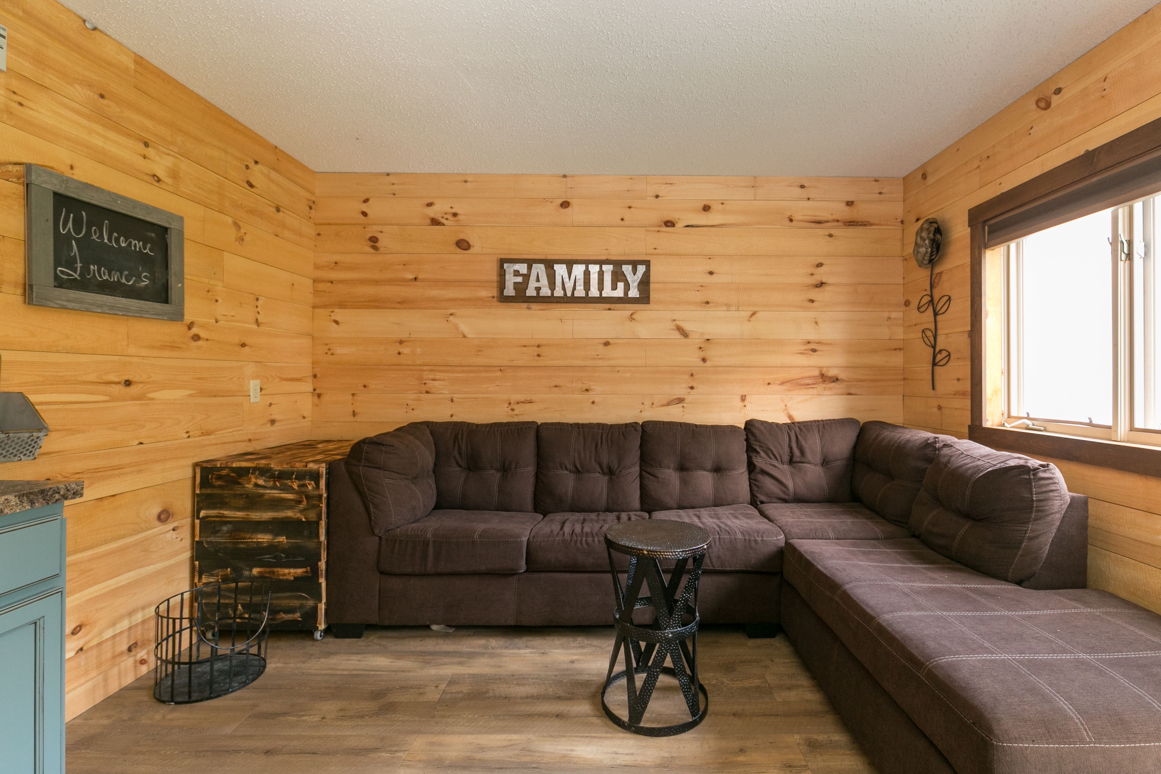 A cozy room with a brown sectional sofa, a small stool, and a chalkboard sign that reads "Welcome".