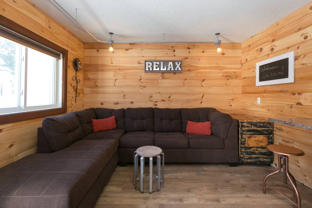 Um quarto aconchegante com um sofá seccional, um banquinho pequeno e uma placa com a inscrição 'RELAX'.