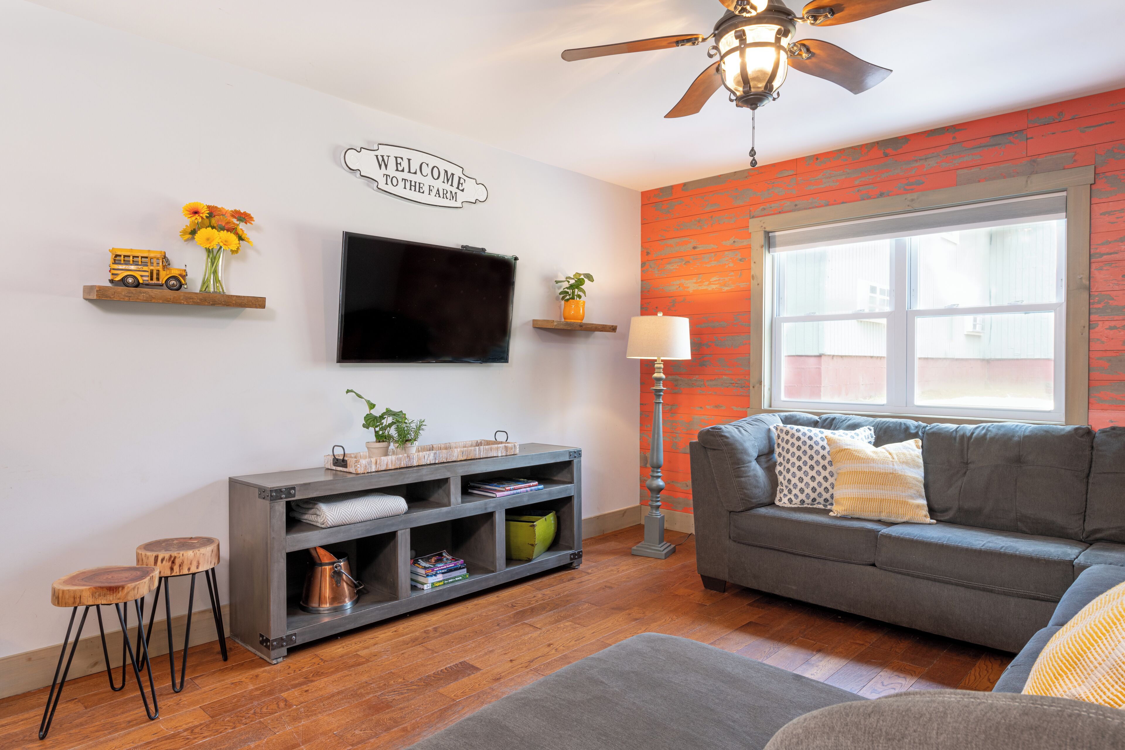 A living room with a grey sofa, a TV mounted on the wall, a bookshelf, a ceiling fan, and a window with a view.
