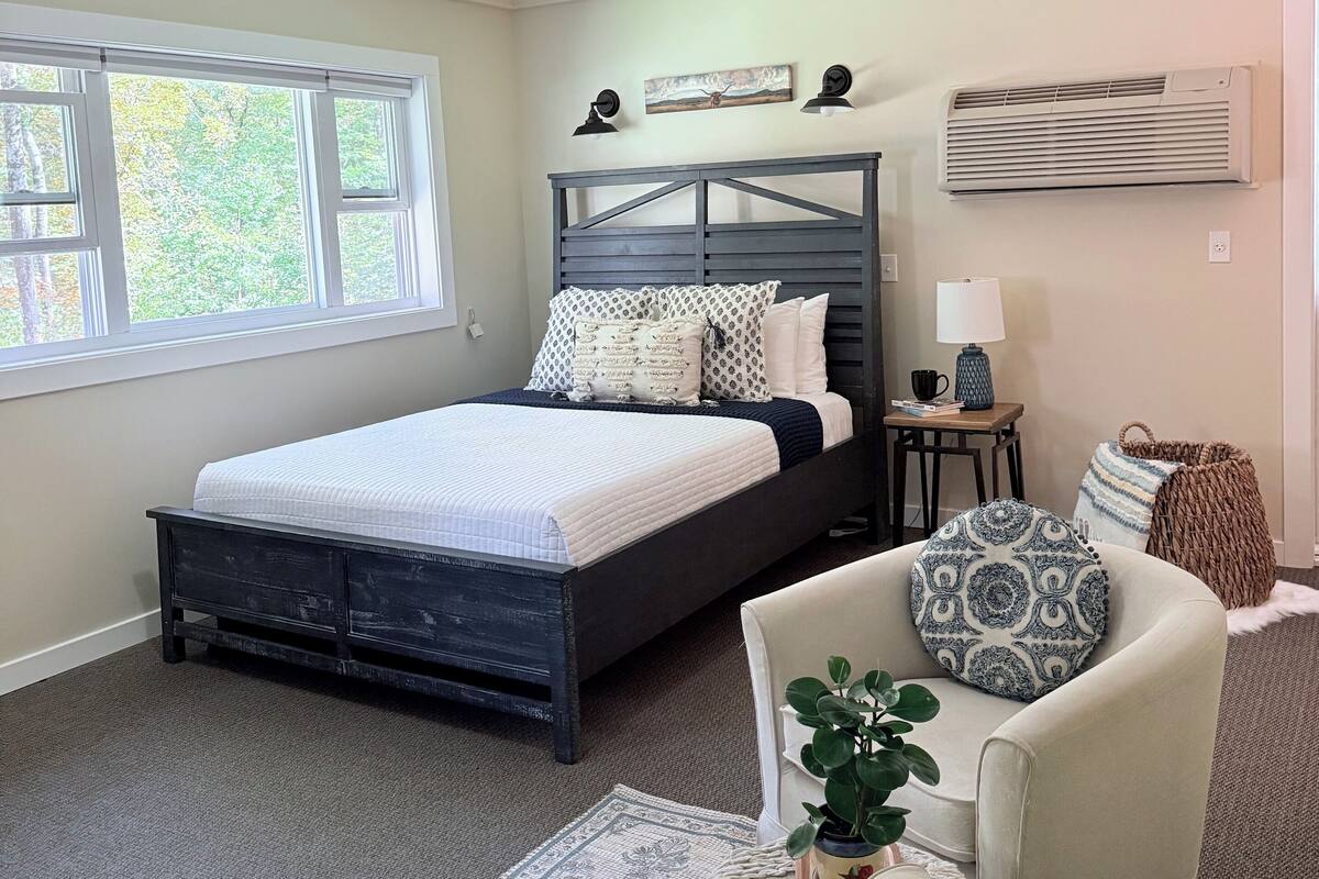 Um quarto com cama, mesas de cabeceira, abajur, cadeira e janela.