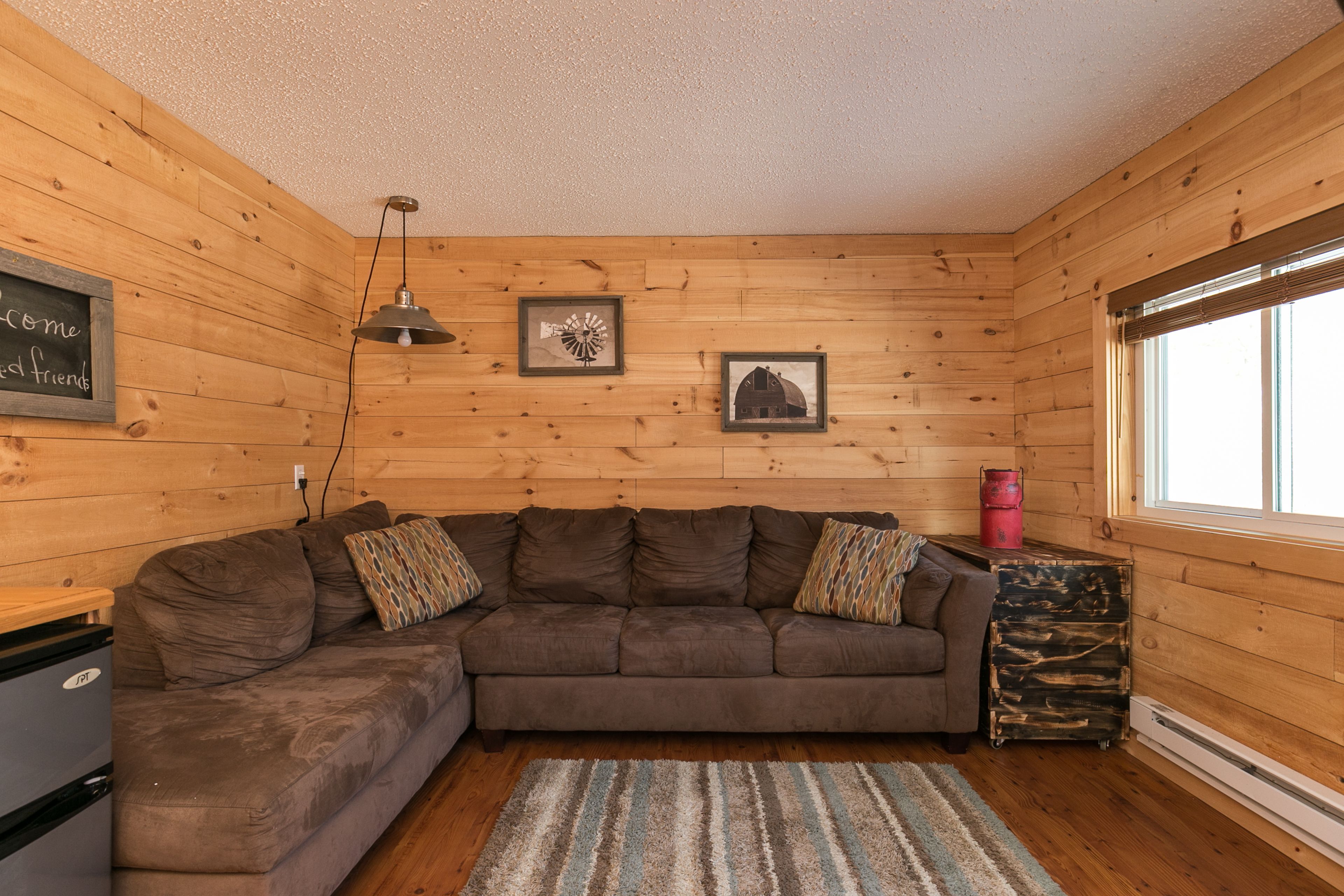 A cozy cabin-style room with a wooden interior, a sectional sofa, a small heater, and a window with blinds.