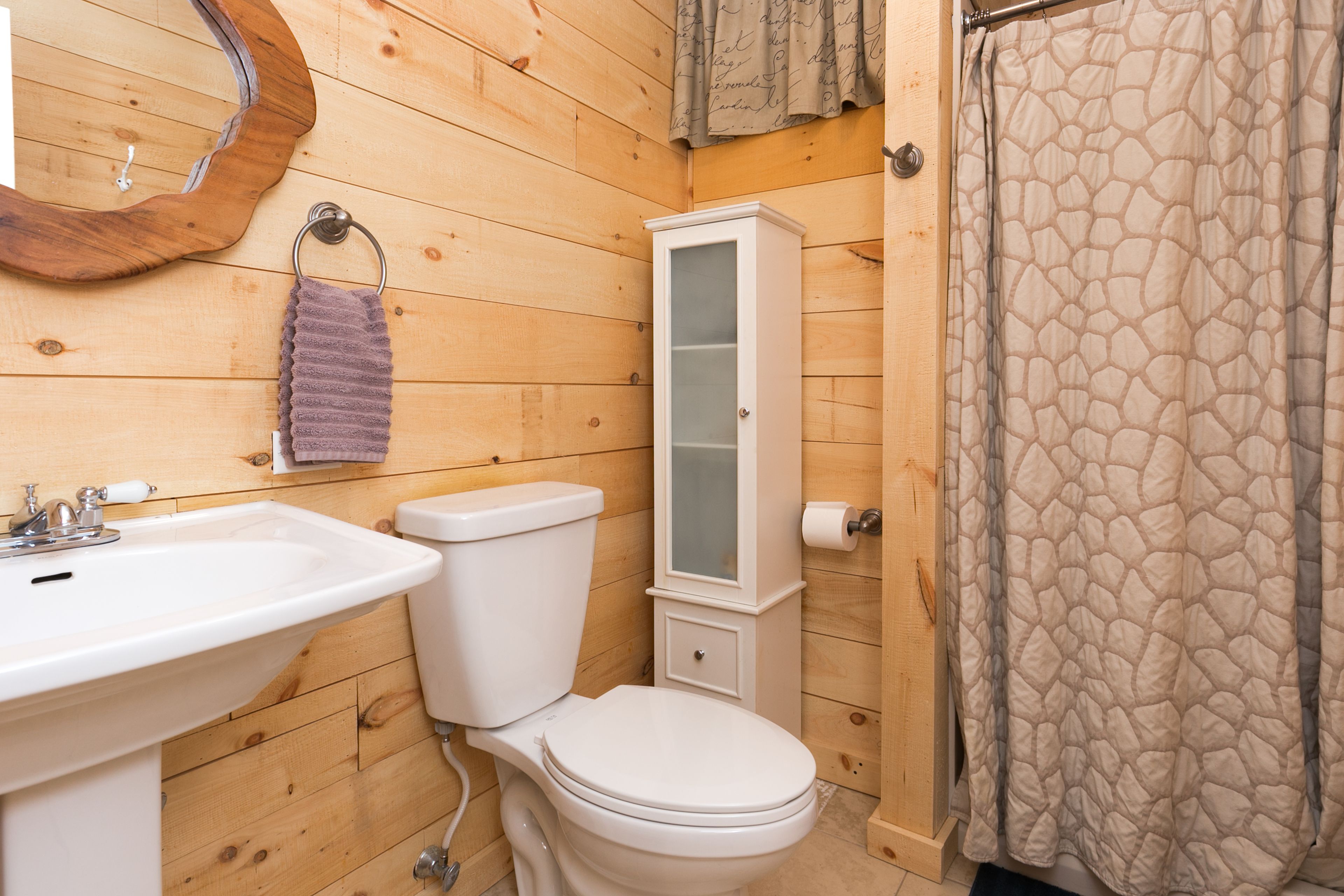 A wooden-paneled bathroom with a white sink, toilet, cabinet, and patterned shower curtain.