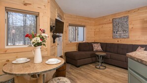 Interior acogedor de una cabaña con detalles de madera, una mesa redonda para comer con platos, un sofá marrón y un taburete pequeño.