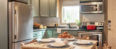 A kitchen with a stainless steel refrigerator, a microwave, and a stove. There is a table with plates and a bowl of food.