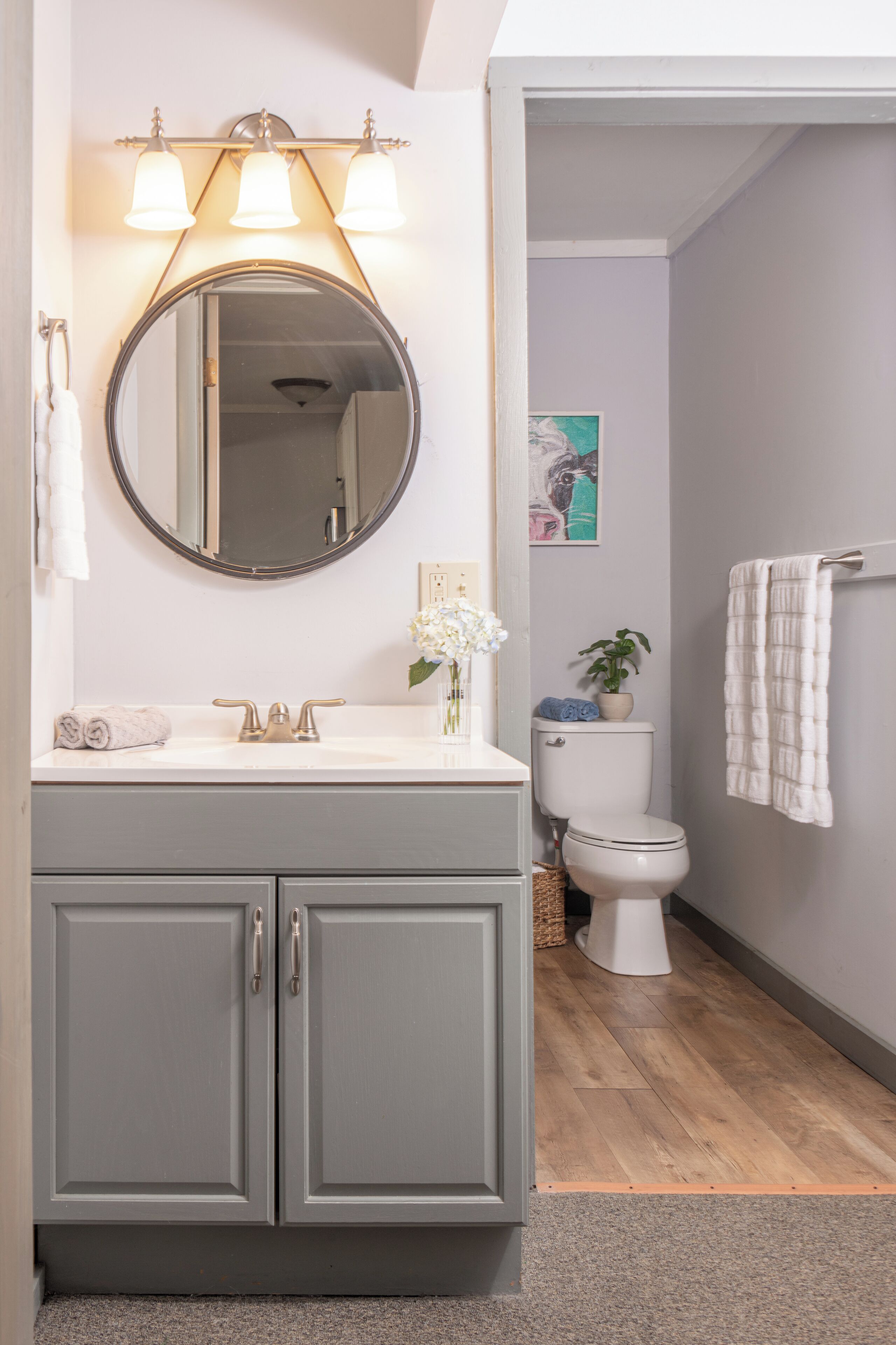 A bathroom with a grey vanity, a round mirror, a white sink, and a toilet.