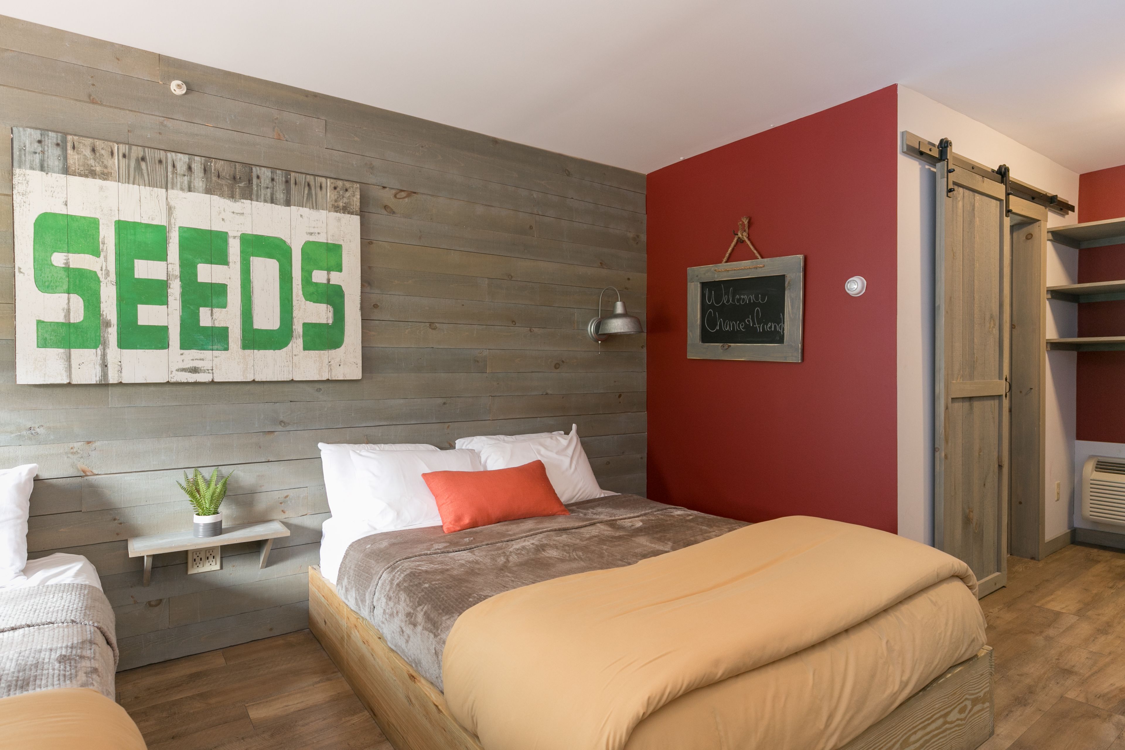 Een slaapkamer met een houten hoofdbord, een bed met beigekleurige en bruine beddengoed, een rode muur en een bordje met de tekst 'SEEDS'.