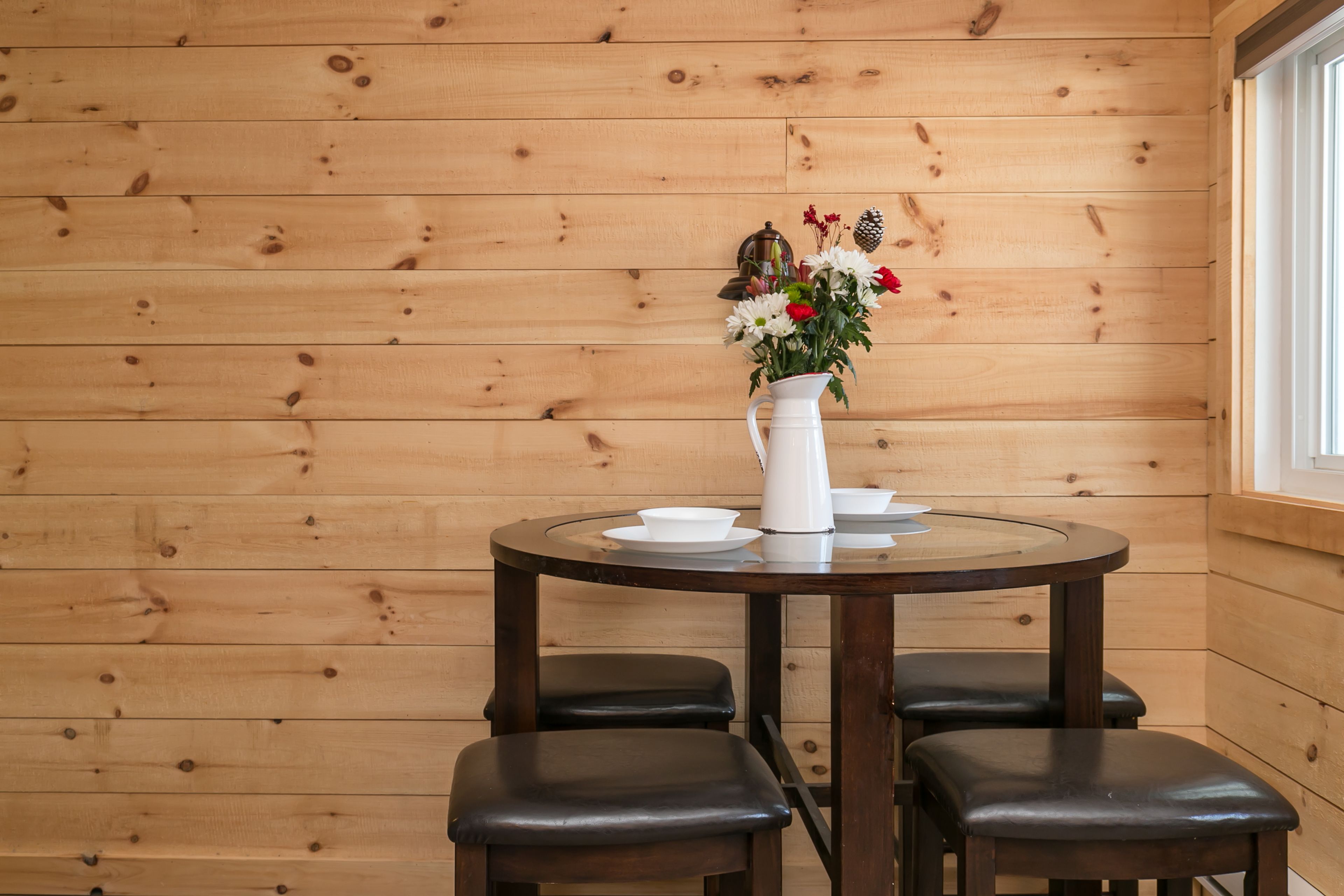 Un intérieur en bois avec une table ronde, quatre chaises, un pichet blanc orné de fleurs et une fenêtre.