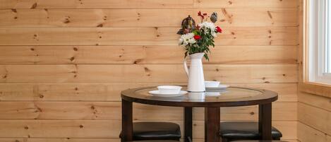 Un intérieur en bois avec une table ronde, quatre chaises, un pichet blanc orné de fleurs et une fenêtre.
