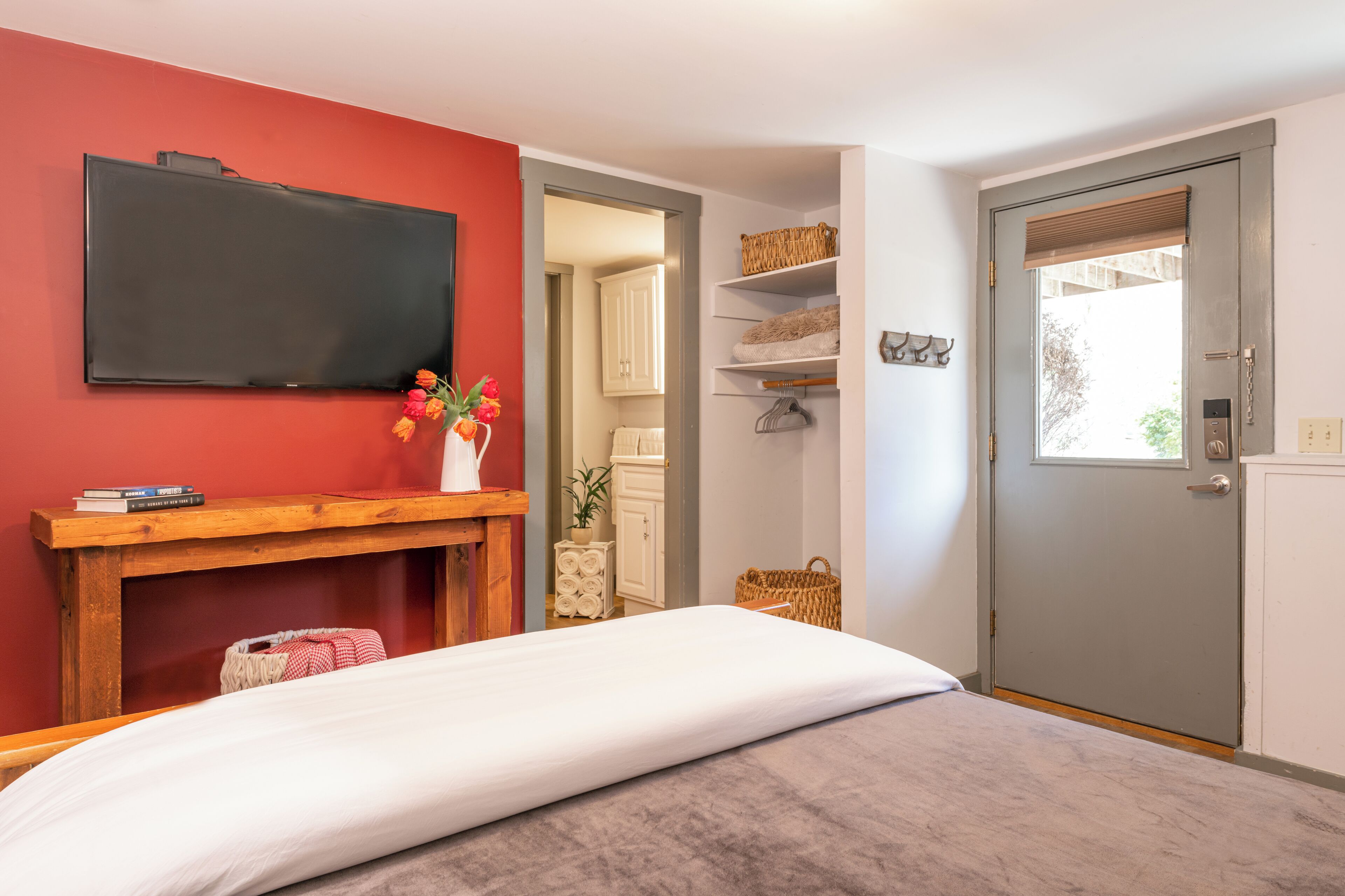 Une chambre à coucher avec un lit, une table de chevet en bois, un téléviseur à écran plat fixé au mur, une porte avec une fenêtre et une petite armoire blanche.