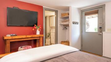 A bedroom with a bed, a wooden bedside table, a flat-screen TV mounted on the wall, a door with a window, and a small white cabinet.
