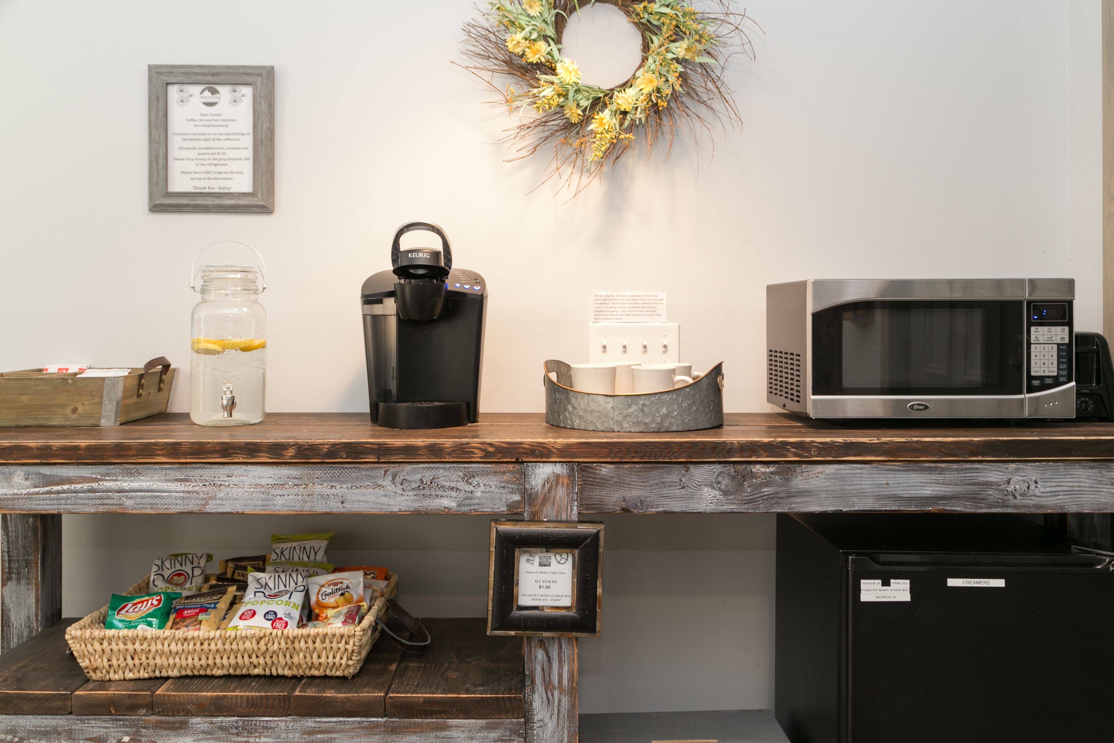A kitchen counter with a coffee maker, a microwave, and a refrigerator. There is a basket of snacks and a jar of lemons on the counter.