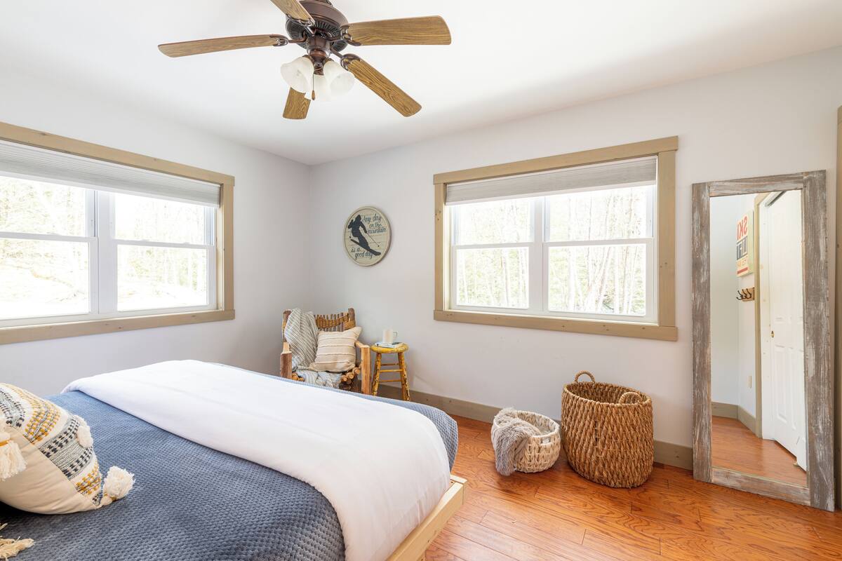 Um quarto com uma cama, um ventilador de teto, duas janelas, uma cadeira, um banquinho e uma porta.