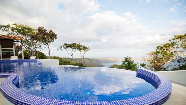 A circular infinity pool with blue mosaic tiles, surrounded by a white pool deck, overlooking a scenic landscape with trees and a distant view of a body of water. - Pacaya Lodge & Spa (Catarina)