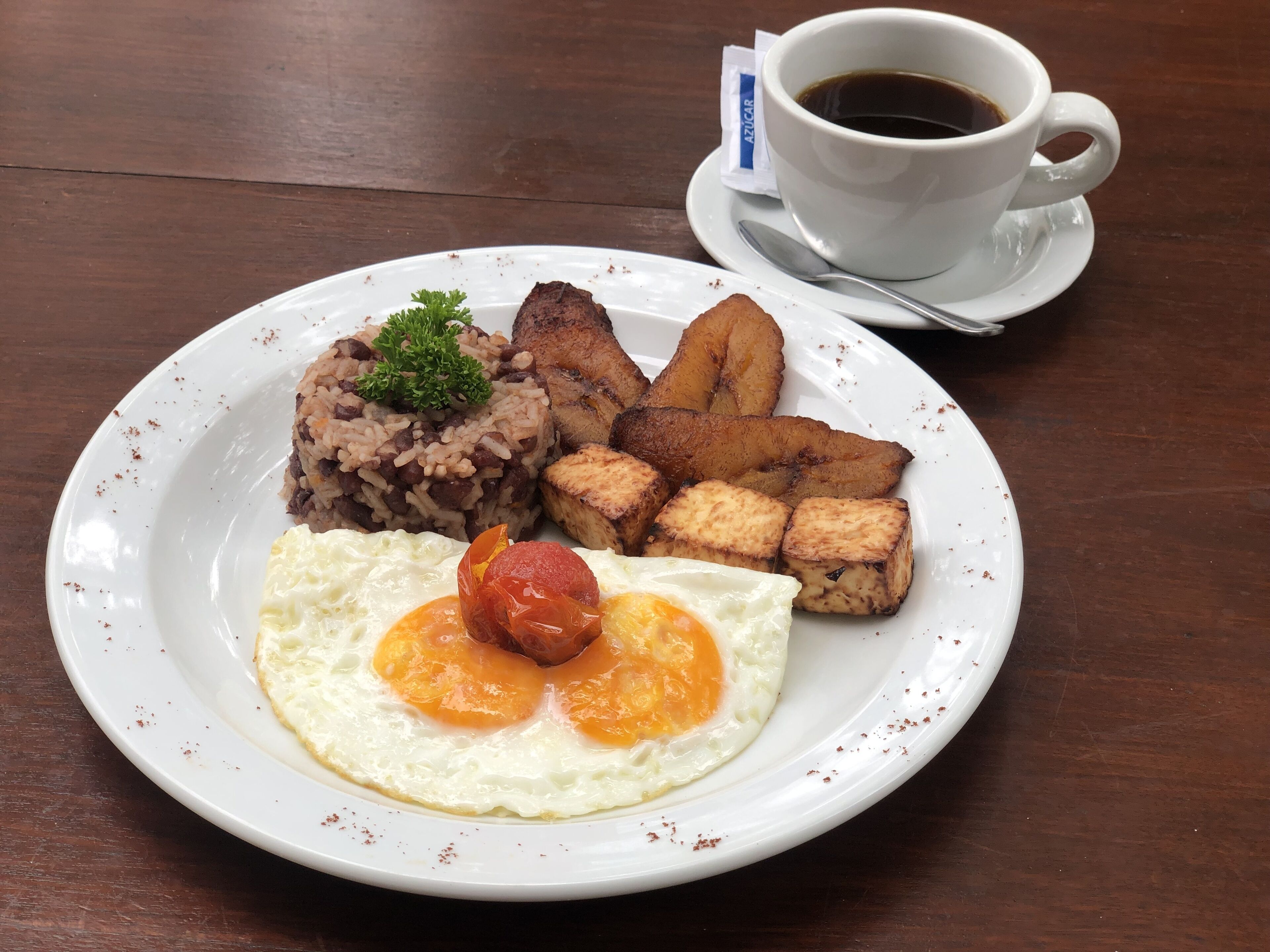A plate with rice, beans, fried egg, and plantains, accompanied by a cup of coffee on a wooden table.