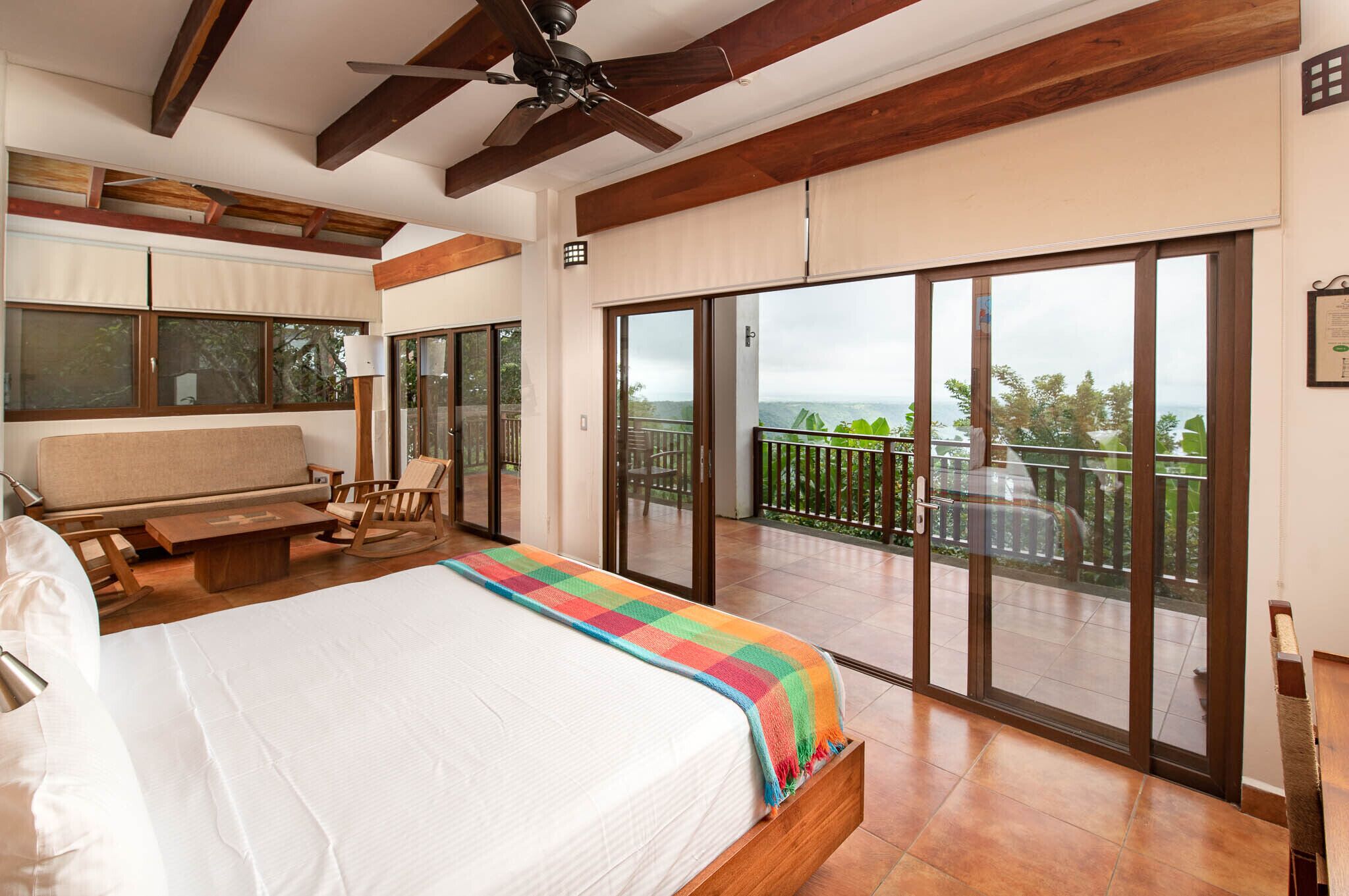 A bedroom with a large bed, a balcony with a view, and a ceiling fan.