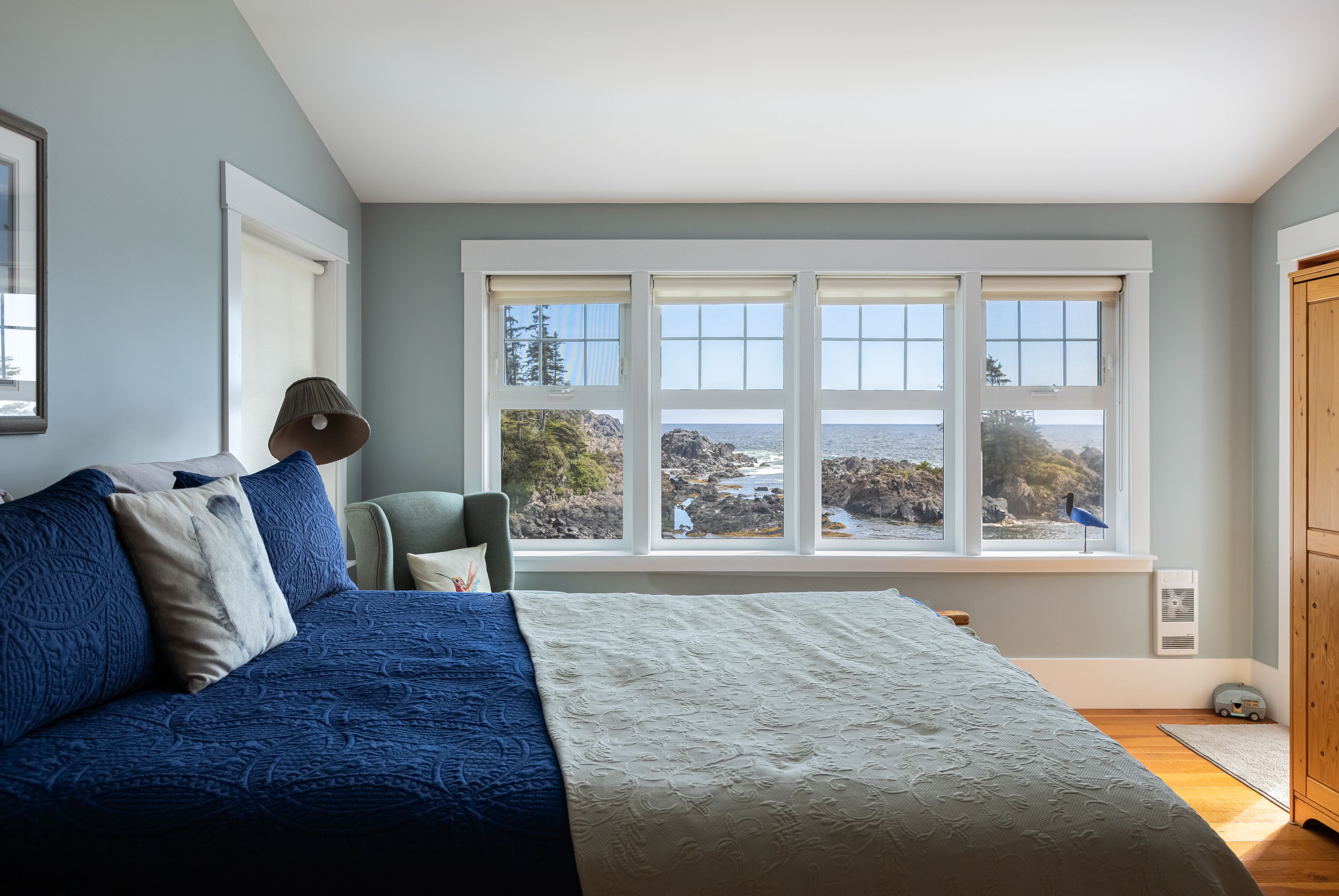 Een slaapkamer met een groot raam, een bed met blauw-witte beddengoed, een stoel en een houten garderobe.