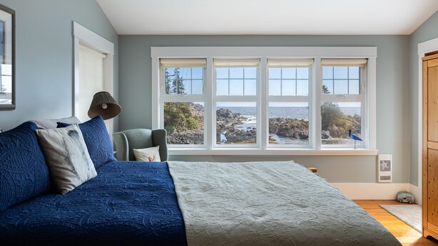 Ein Schlafzimmer mit großem Fenster, einem Bett mit blau-weißer Bettwäsche, einem Sessel und einem Holzschrank.