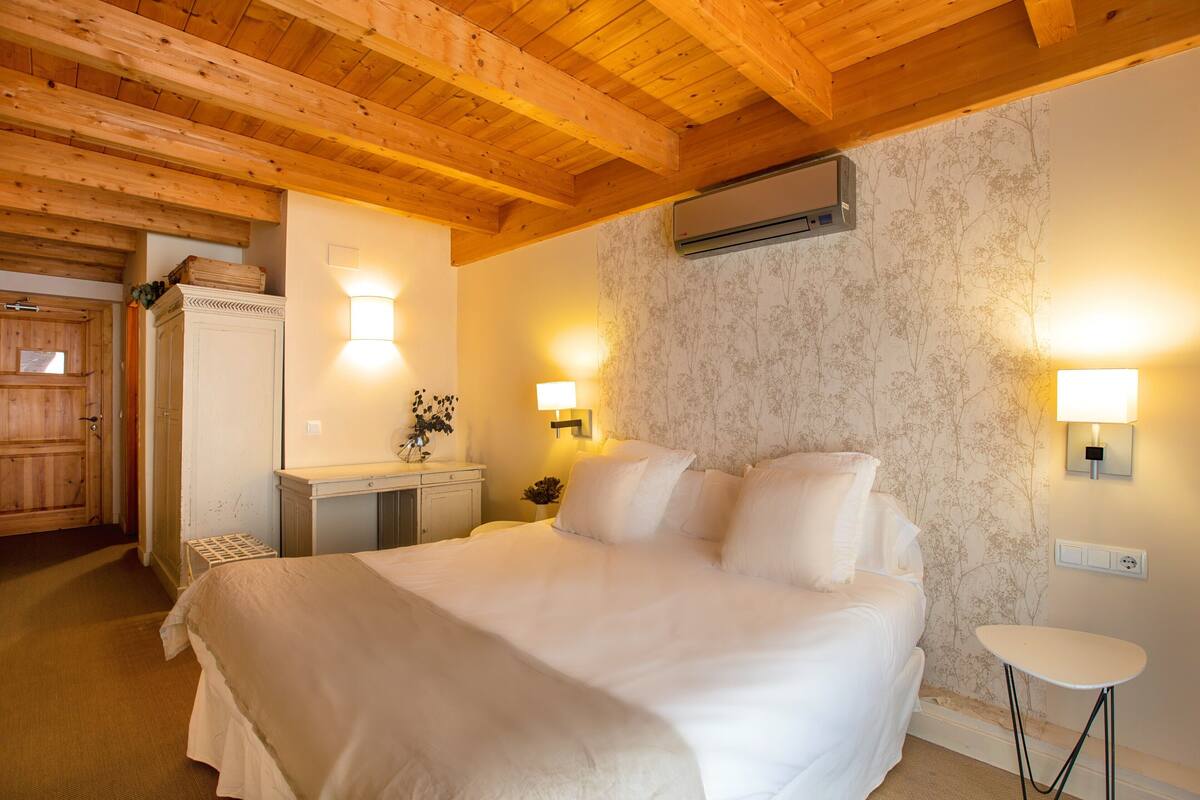 A bedroom with a wooden ceiling, a bed with white bedding, a small bedside table, and two wall-mounted lamps.