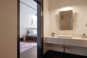 A modern bathroom with a large mirror, white sink, and a bedside table with a lamp. - Hotel Hardware (Dayton)