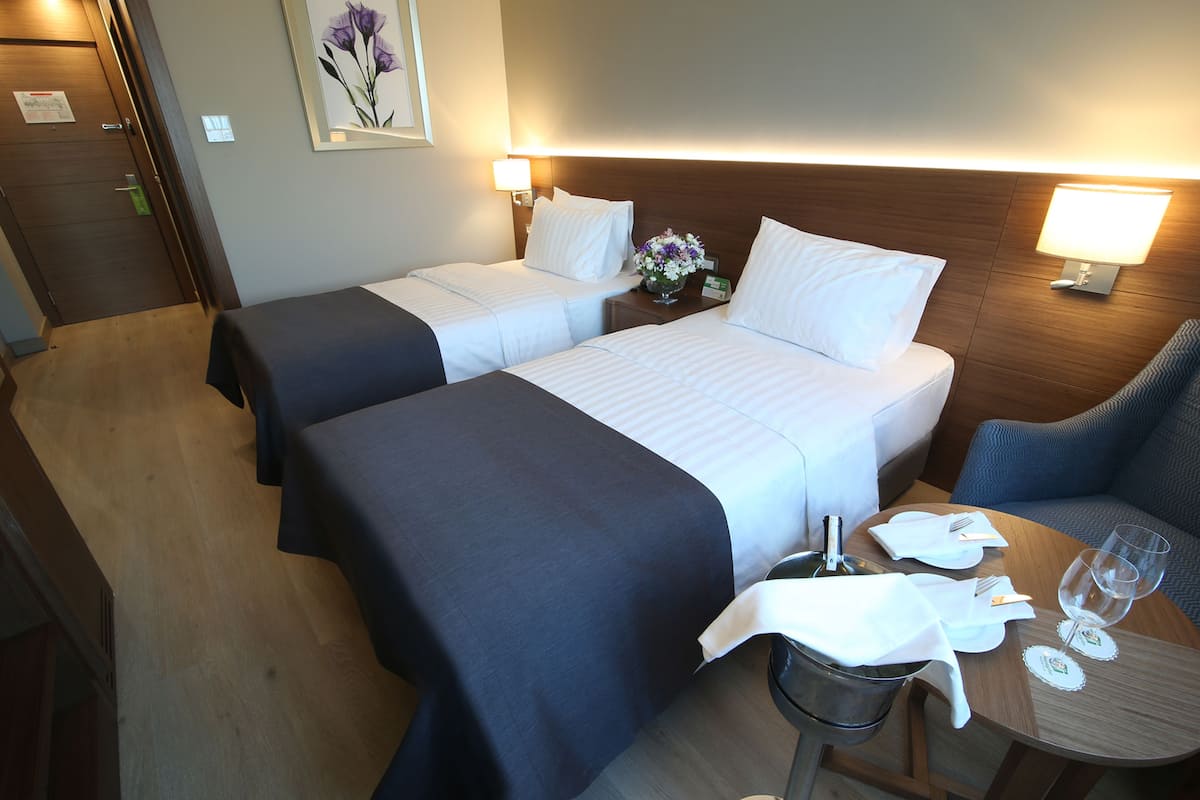 Ein Hotelzimmer mit zwei Betten, einem Esstisch mit Stuhl, einem kleinen Tisch mit Glas und Servette sowie einer Wandlampe.