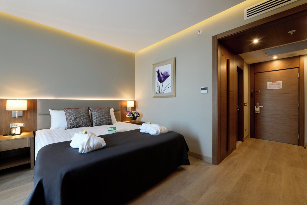 Une chambre d’hôtel avec un grand lit, des lampes de chevet, une table de nuit, un tableau encadré au mur et une porte en bois.