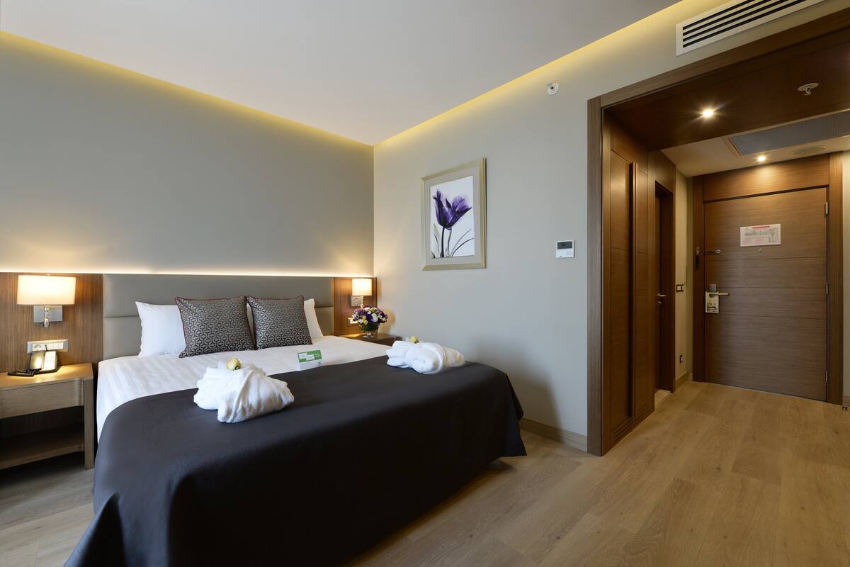Ein Hotelzimmer mit einem großen Bett, Nachttischlampen, einem Nachttisch, einem gerahmten Bild an der Wand und einer Holztür.